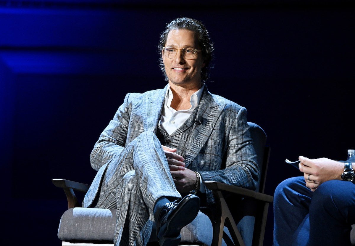 Matthew McConaughey smiles dressed in a grey suit sitting on a grey chair